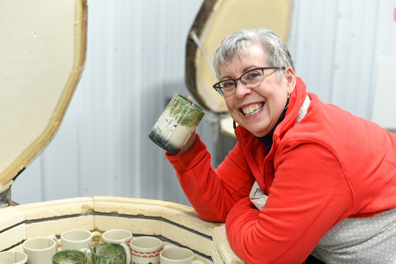 A photo of Canadian potter Susan Robertson holding a mug with birch trees. The mug is fresh from the kiln and she is leaning on the edge of the kiln. She is smiling and facing the camera.